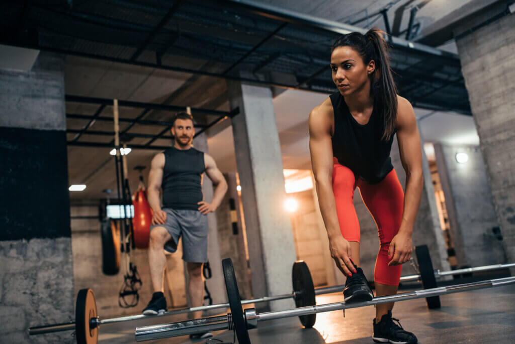 Pareja se prepara para hacer encogimiento de hombros con barra en el gimnasio.